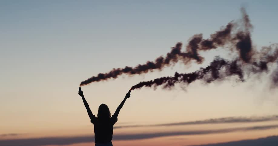 Girl holding smoke flares high over her head and the smoke is leaving a long trail in the air