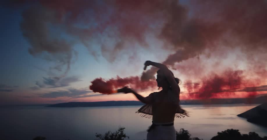 A woman is seen dancing with 2 objects that emit smoke on a shore