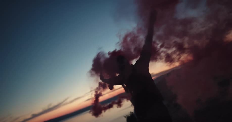 Silhouette of young couple dancing and jumping with smoke flares in front of a lake at sunset