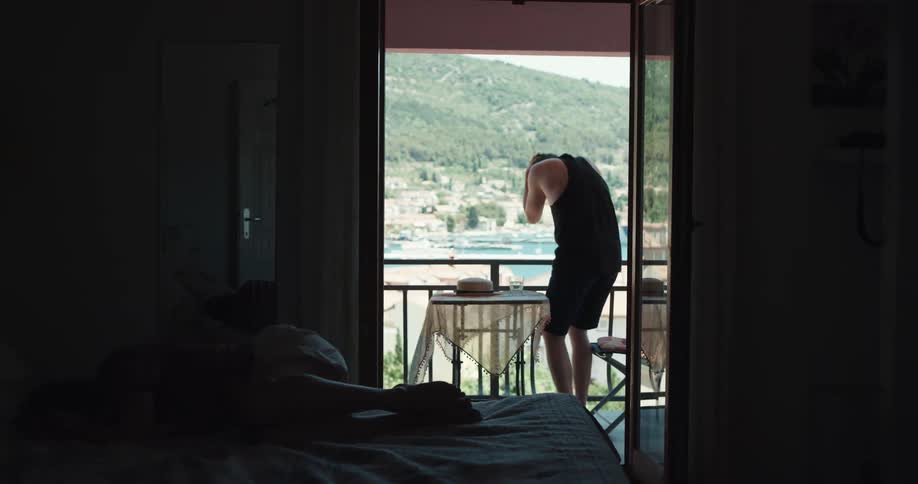 Sad man with black tank top sits down in chair in the balcony after fighting woman lying down on bed crying