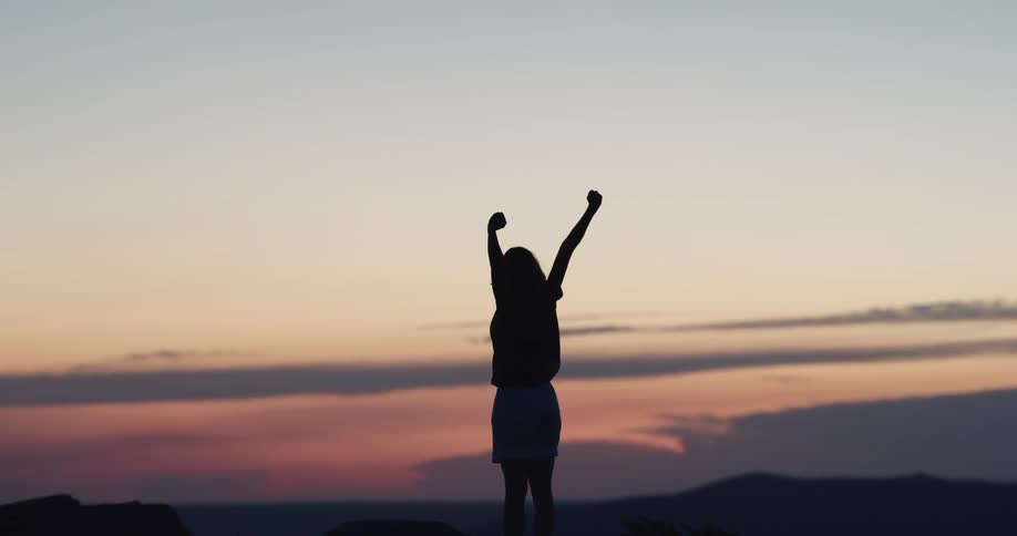 Woman stretches her arms in the air as she faces the distant mountains at dawn