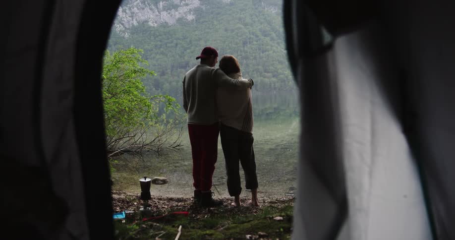 A female camper stretches her back and arms before being handed a cup of coffee and being embraced by a male camper who comes to stand beside her