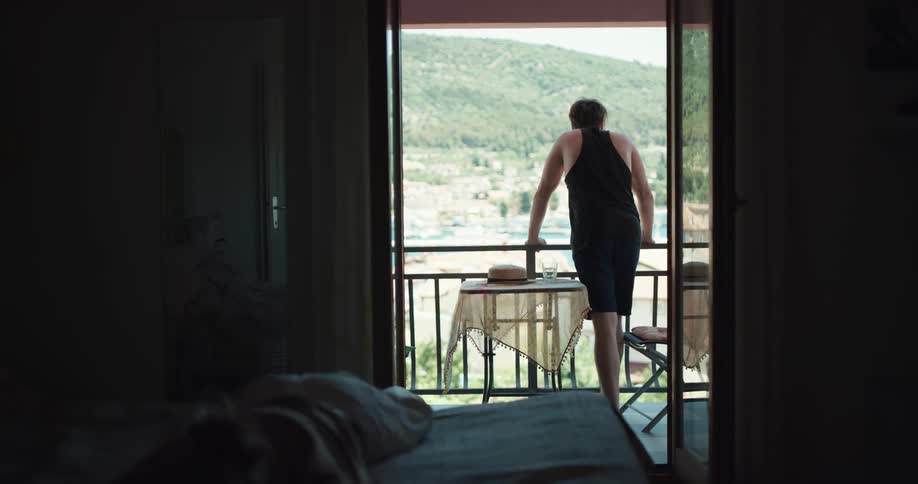 A woman cries in bed as her partner stands on the balcony of their hotel room overlooking the ocean