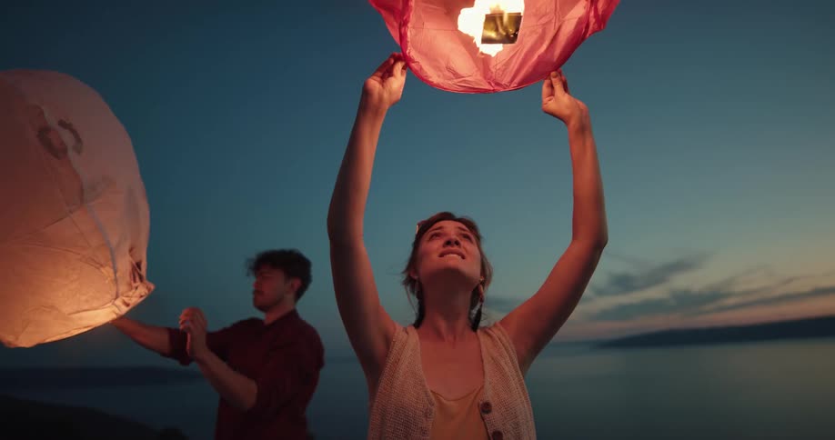 A man and woman stand on the shore and hold paper lanterns with fire burning in them