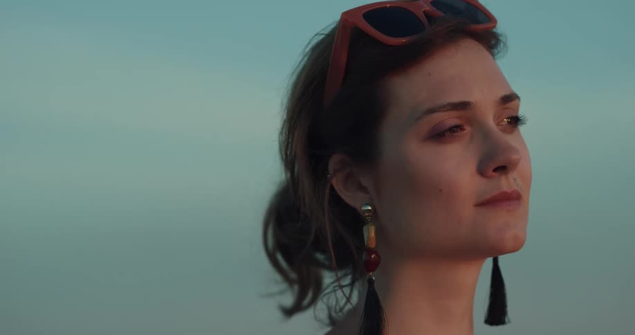 Woman stands looking as light brightens her features then closes her eyes against blue of sky