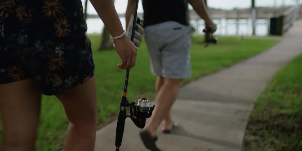 A man and a woman are walking towards a water body while holding fishing rods