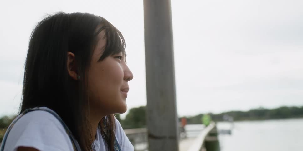 A young woman relaxes against the railing of a wooden pier on a lake on a cloudy day