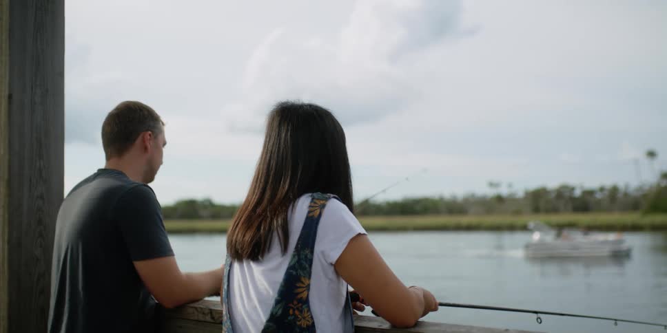 Young couple fishing from a bridge looking at a boat on a lake