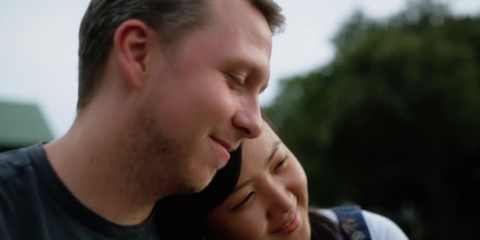 A woman laying her head on a man shoulder with trees the sky and clouds in the background