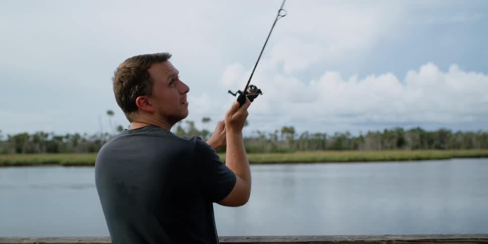 Man leisurely fishing on a pier near a lake while a boat passes by