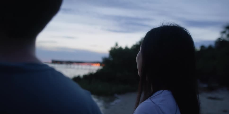 A man and woman standing by a lake with trees the sky and clouds in the background