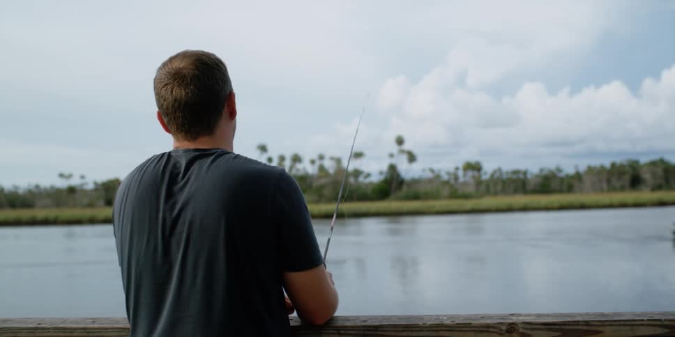 A man standing on a dock by a river fishing with a fishing rod with trees the sky and clouds in the background