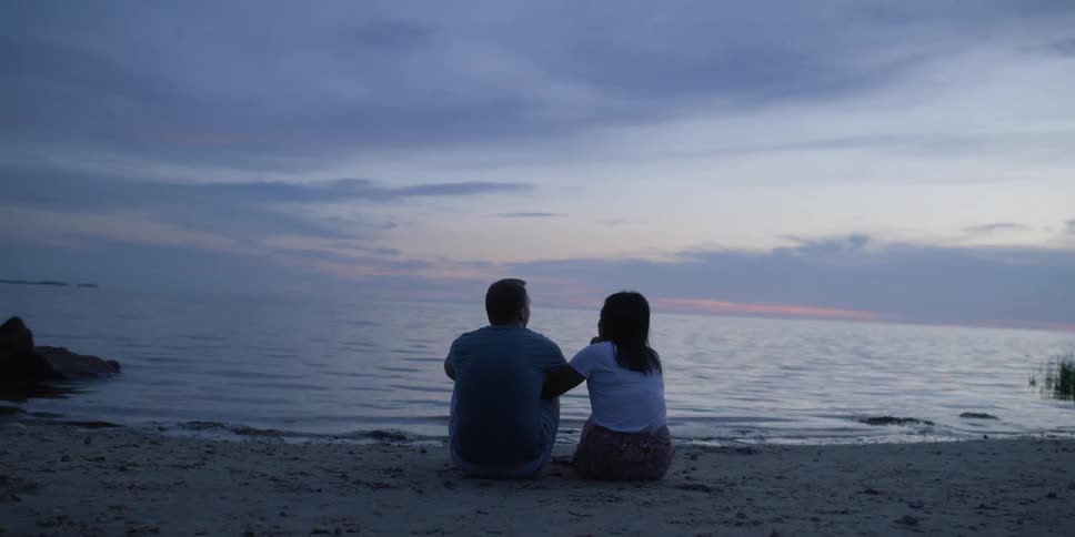 A man and a woman sit next to one another on a beach in front of water