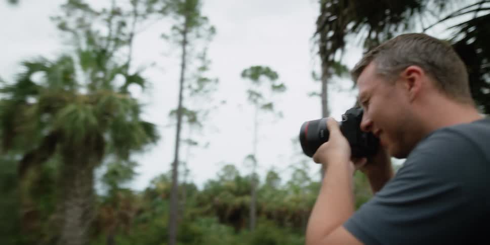 A man with a camera taking a picture of a woman on a walkway with trees the sky and clouds in the background