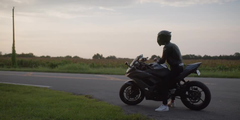 A motorcyclist sits contemplatively atop his motorcycle parked on the side of a road through a field in the evening