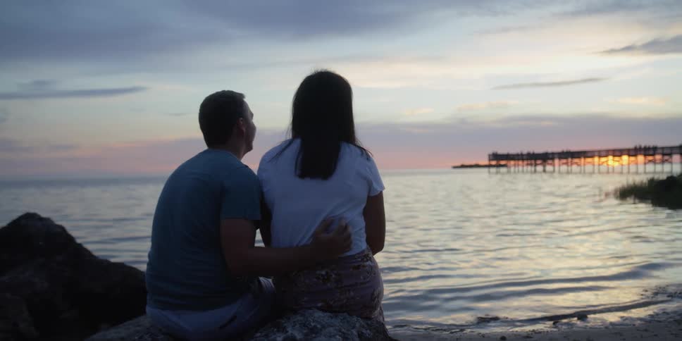A man and woman watch the setting sun over the ocean while sitting together on a large rock on the beach
