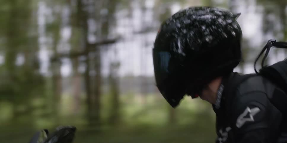 A motorcyclist wearing a backpack rides along a road through a woodland during the day