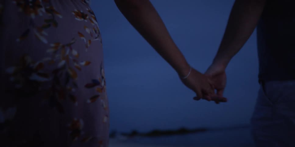 A man and woman hold hands while walking along a beach at night