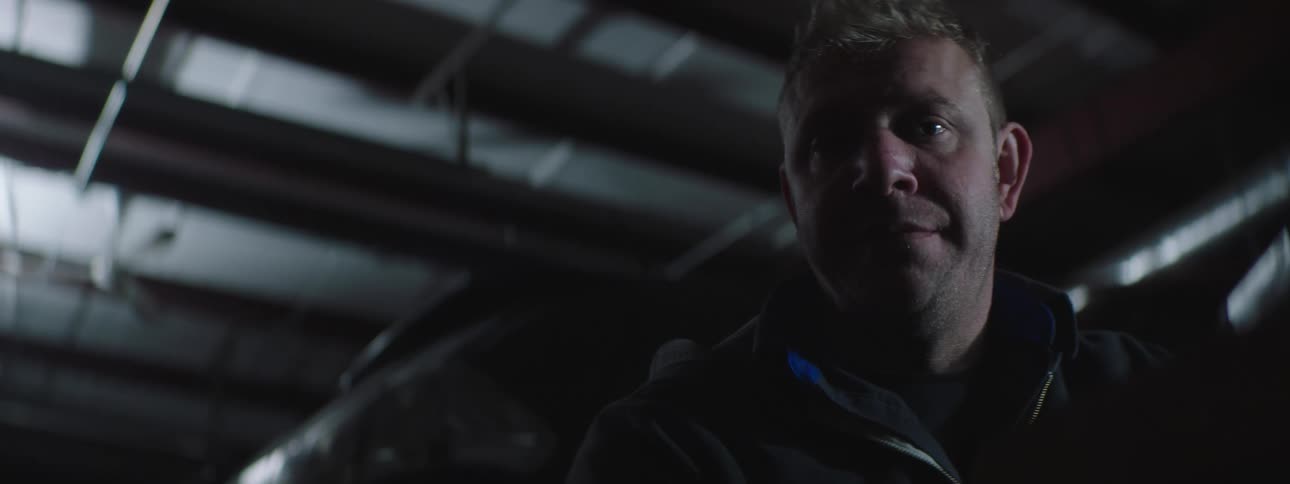 A man standing in a dimly lit metal building turns his head to look into the camera