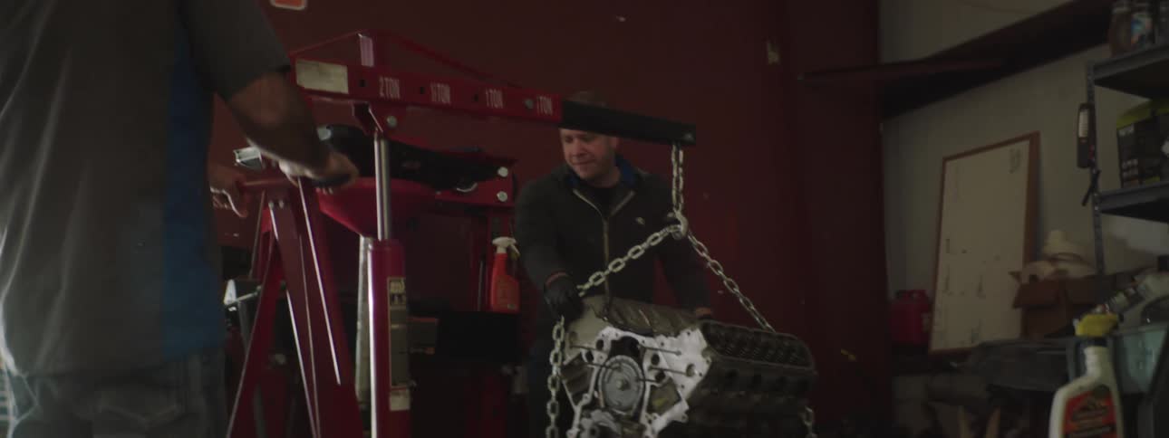 Two men slowly move a car engine in a garage using an engine hoist