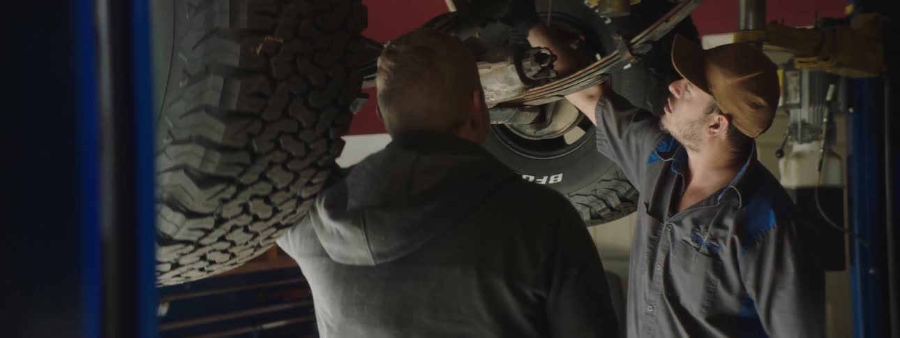 Two men stand underneath a vehicle thats lifted in the air in a commercial garage