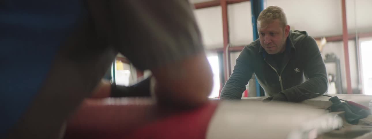 Man wearing a sweater with a zipper talks to another man across from him in a workshop