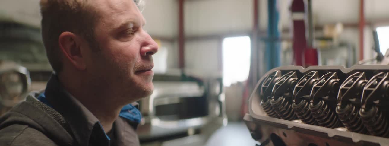 Man touches a vehicle engine with a gloved hand then places a tool on top of it