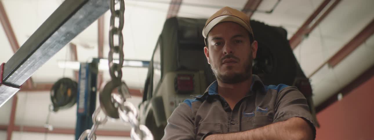 A man with a hat in a garage staring with a vehicle and a wall in the background
