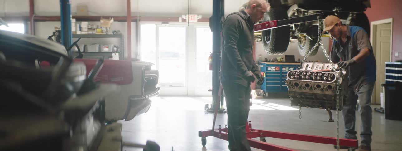 Two men approach a car part hanging from chain and examine it in a garage