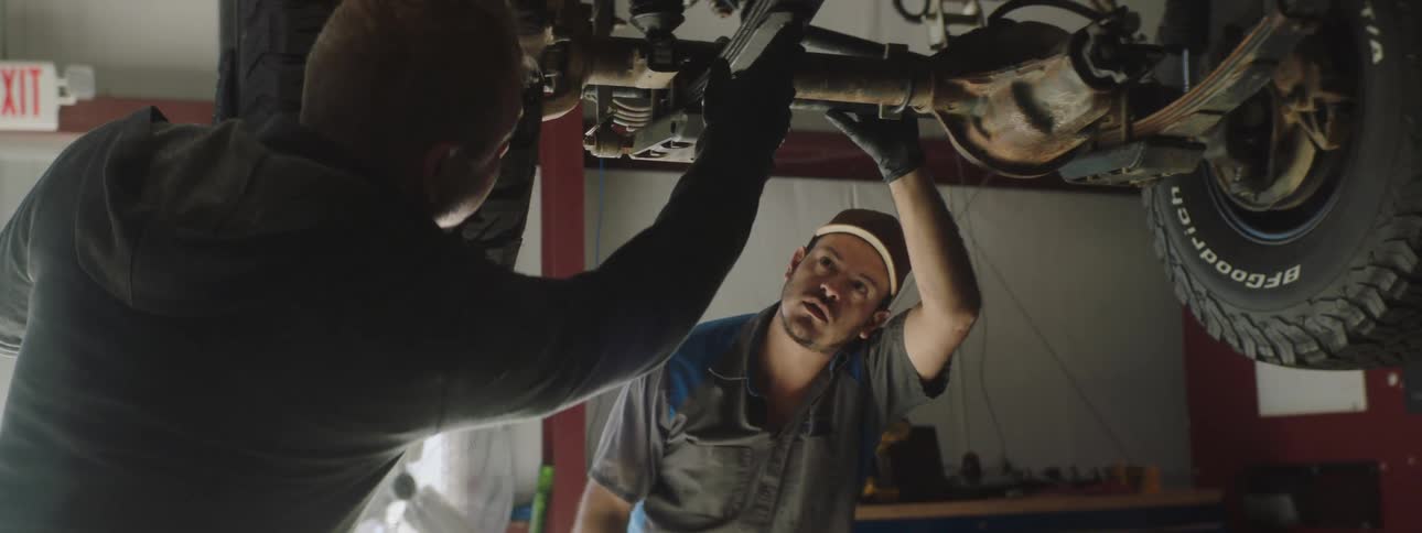 A pair of men examine the bottom of a suspended car in a garage
