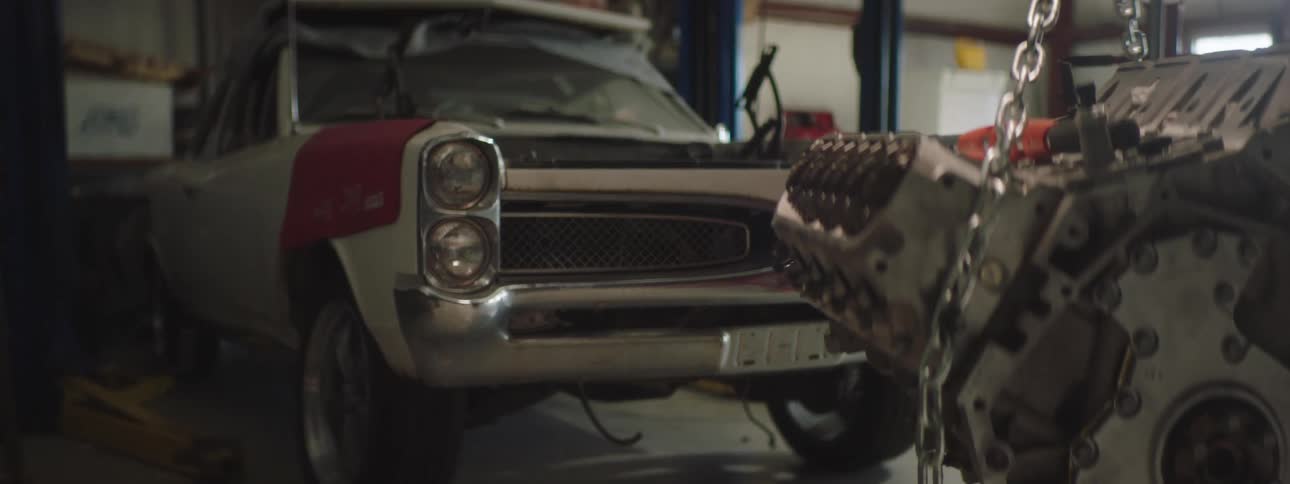 A garage with an engine hanging on chains with a vehicle and a wall in the background