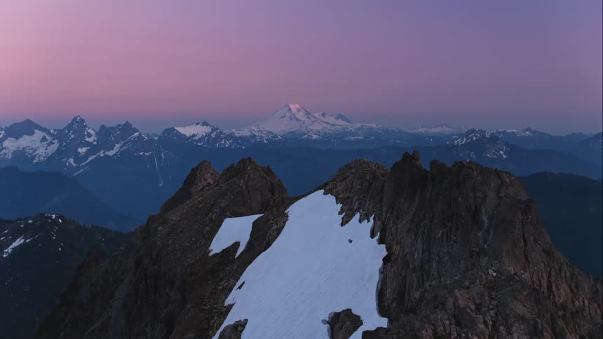 Aerial push through mountain peak to reveal snow capped mountain range in distance at dusk