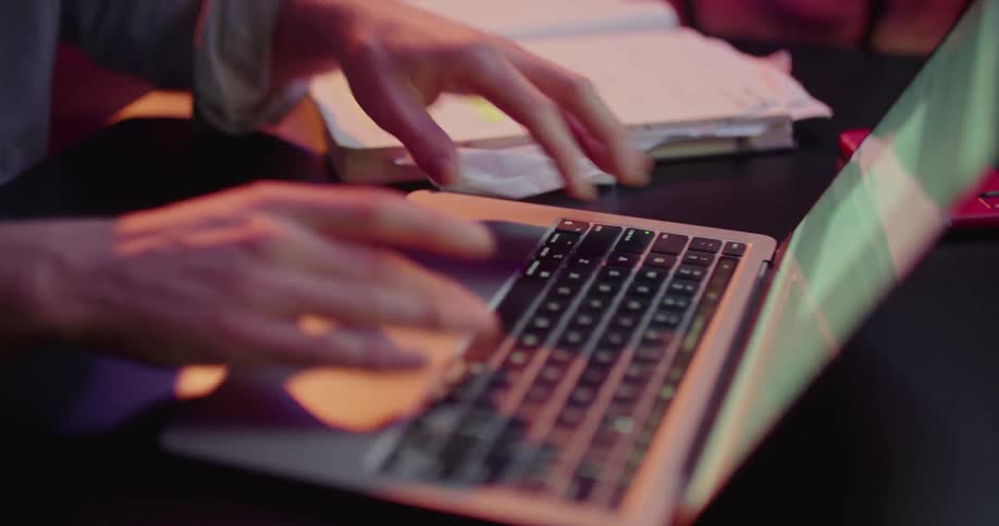 A male flips through pages of his notebook before typing some numbers into his laptop