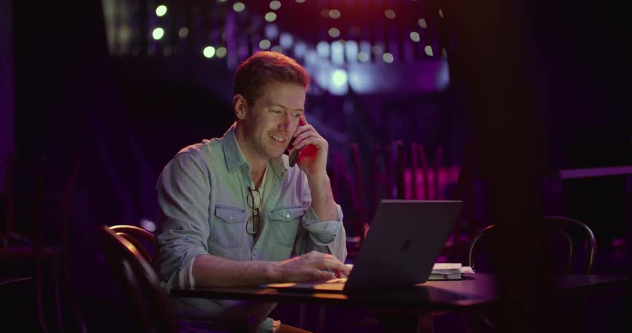 A man sits in a dark room and answers his cell phone and talks while using his laptop