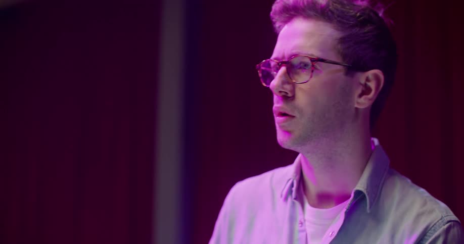 A man typing on laptop under a purple light indoors looks up for a moment to collect his thoughts