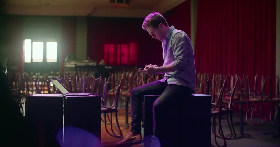 A man leans against a large speaker working on a laptop and then dialing on his cell phone
