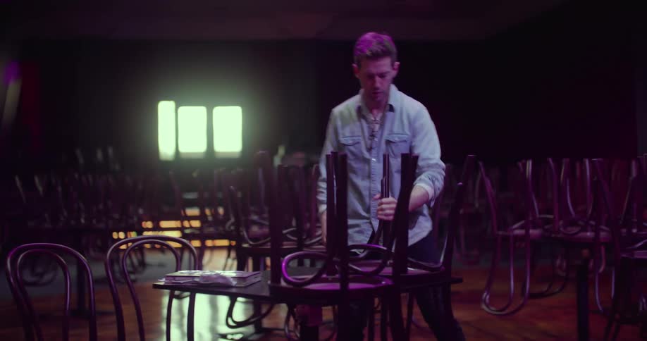 A man in a nightclub taking chairs from on top of the table and placing them on the floor with a wall and window in the background