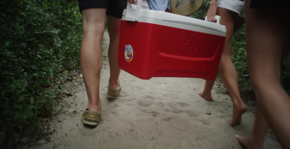 Several people walking a sandy trail carrying a cooler and a surfboard