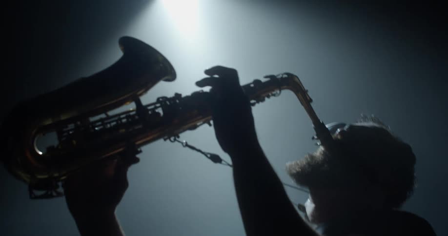 Man with beard plays saxophone on dim smoky stage partly silhouetted by spotlight