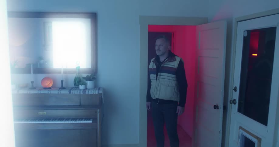 Man walks rigidly from hallway into room standing frozen facing glowing rectangular column