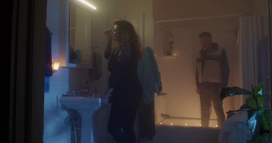 A woman applies her make up in a home bathroom with a man watching from the shower
