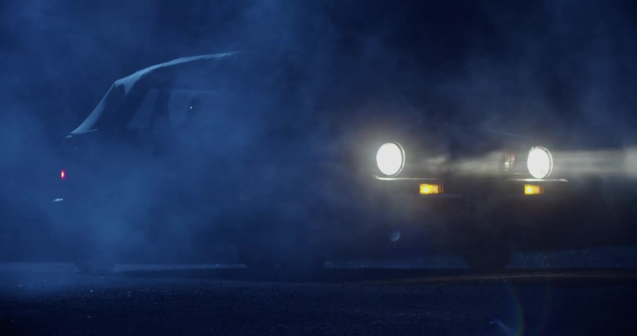 A small car is idle on a dark and foggy road while its headlights are on