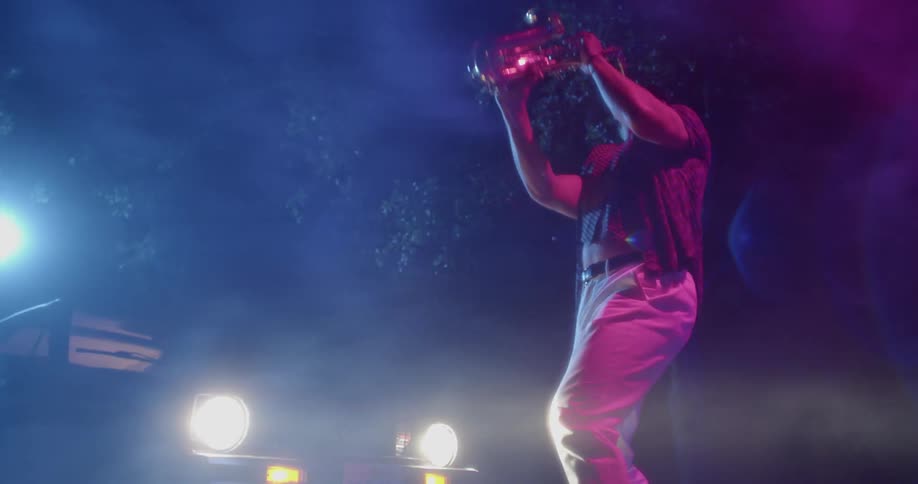 Man plays a saxophone outdoors as a cars headlights illuminate him