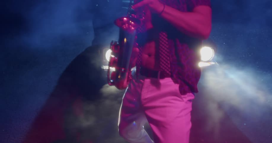 A man stands outside at night in front of a stationary car while he plays saxophone