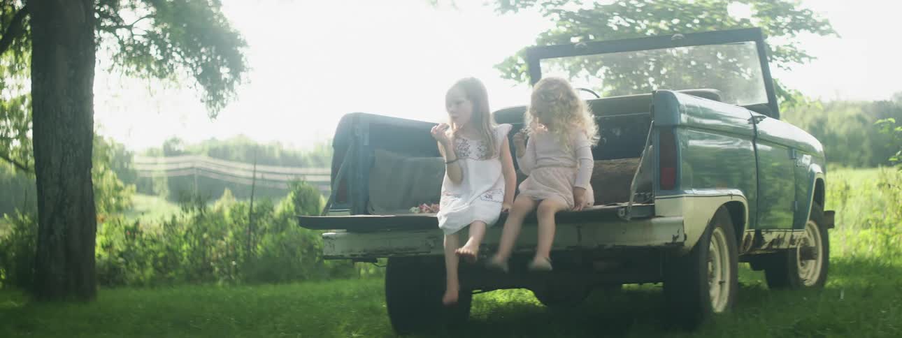 Two little girls sit on a truck tailgate swinging their legs and eating on a bright sunny day