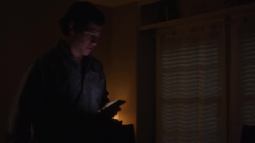 Man works on a laptop then walks across the room carrying a cellphone as lights flash outside the window