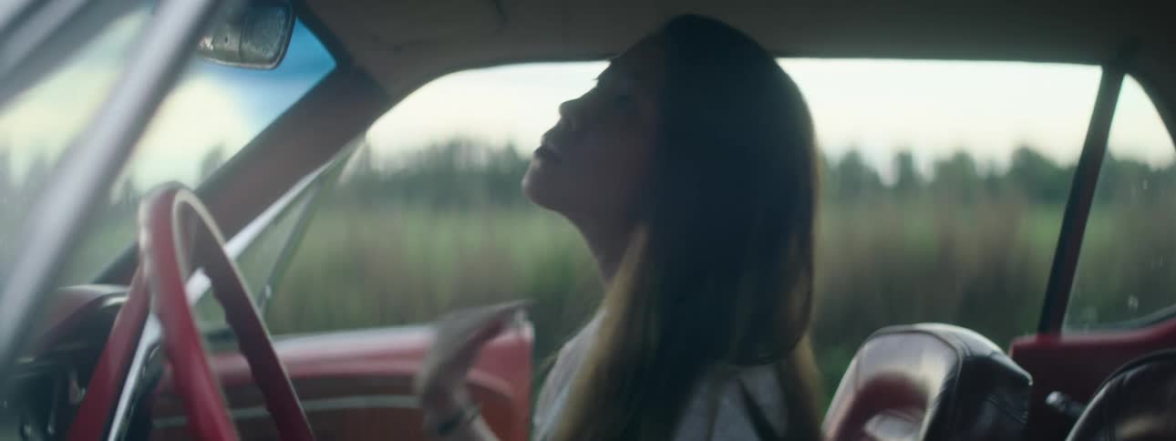 Woman sitting in passenger seat of car combs back her hair before looking out open door toward nearby field