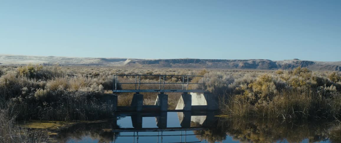 A bridge over a small dam with low mountains in the background under a clear blue sky