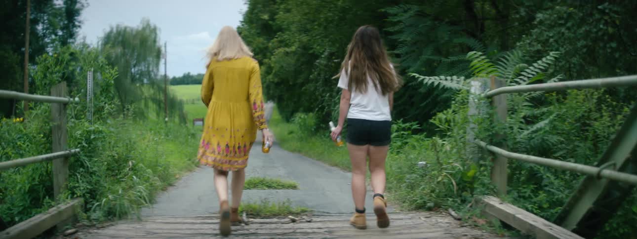 Two women each carrying a bottle walk across a wooden bridge and onto a path that leads past the trees