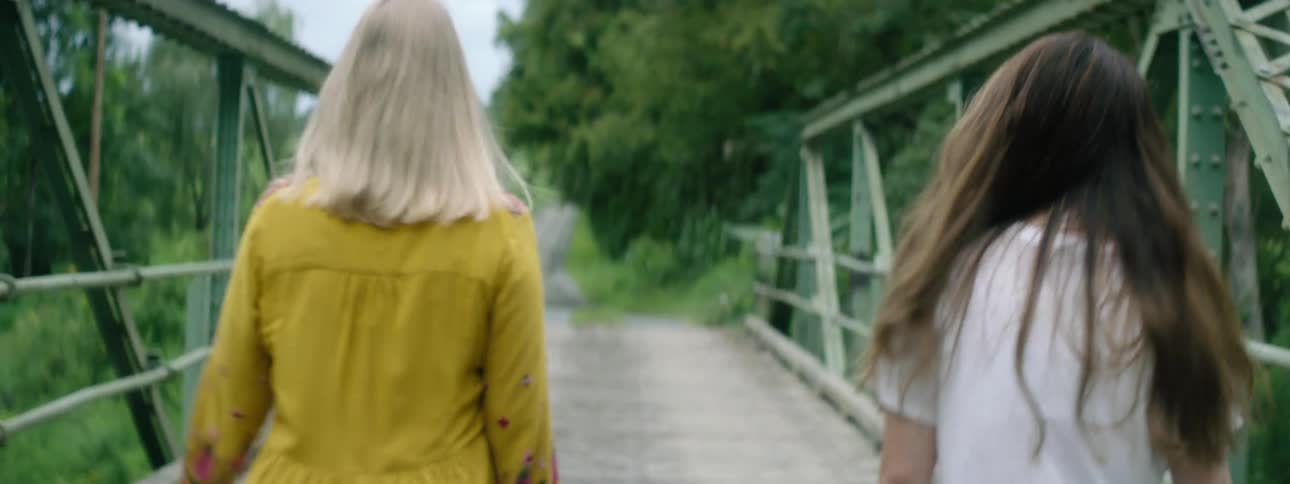 Two women walk over a bridge towards a dirt road next to the trees